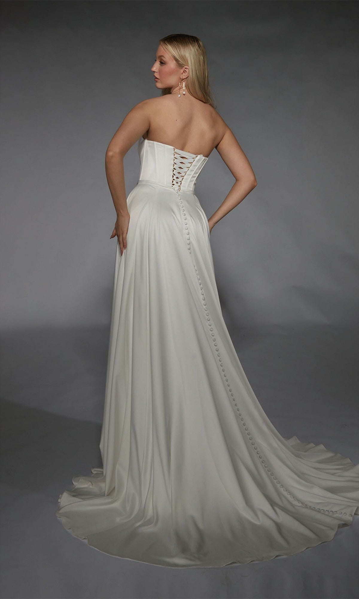 A woman faces away from the camera against a gray backdrop, wearing the Alyce 7111 long white dress—a strapless satin gown with an A-line skirt, lace-up back, and button details.